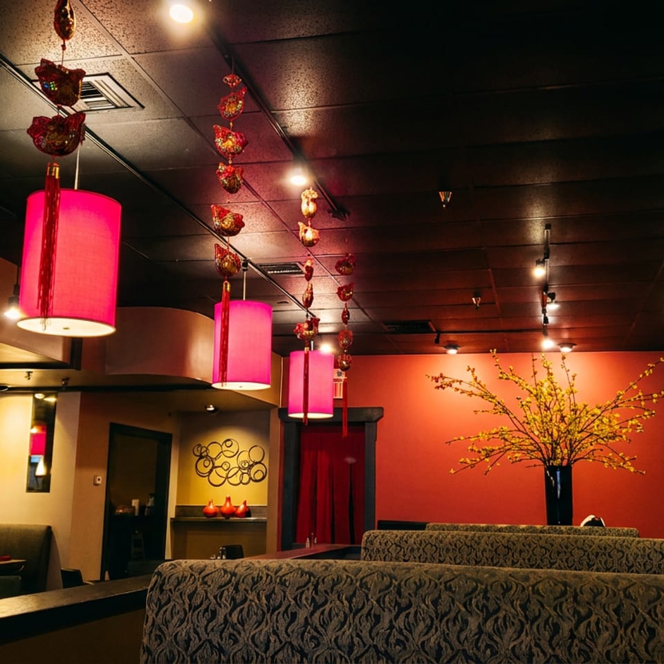 Warmly lit Asian restaurant interior with red lanterns, pink pendant lights, patterned booths, and flowering branches.