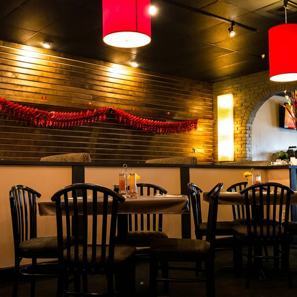 Warmly lit restaurant dining room with set tables, black chairs, red pendant lights, and festive red garlands on a wood-paneled wall.