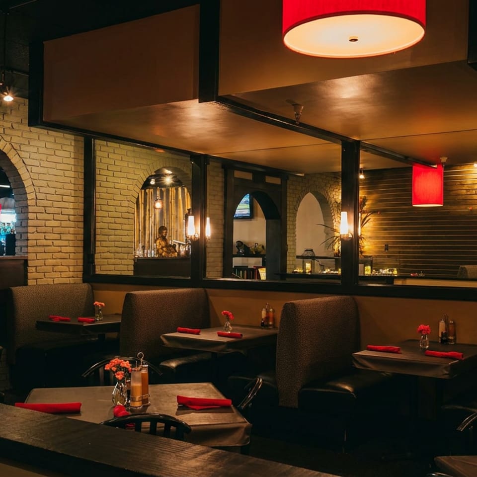 Warmly lit restaurant dining room with red pendant lamps, brick arches, upholstered booths, set tables, and a visible bar.