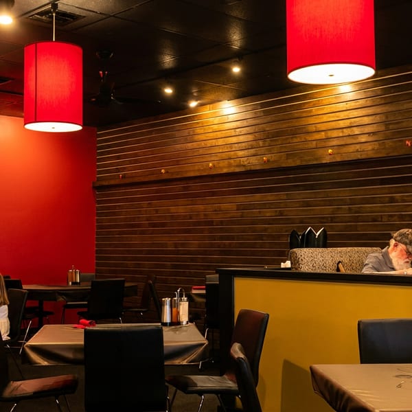 Warmly lit restaurant dining room with red pendant lights, wood-paneled walls, booths, tables, and several diners.