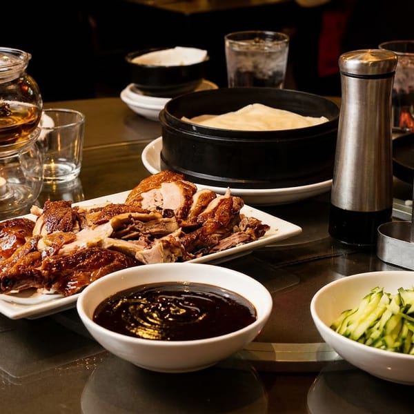 Chinese-style roasted duck with pancakes, dipping sauce, cucumber, and scallions arranged on a restaurant table.