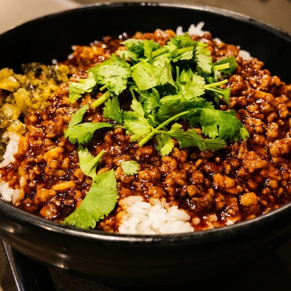 Black bowl of white rice topped with saucy minced meat, cilantro, and sautéed vegetables on a restaurant table.