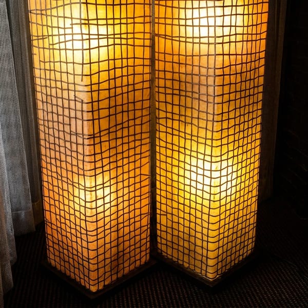 Two glowing amber floor lamps with gridded shades beside sheer curtains and an ornate gold console in a dim interior.