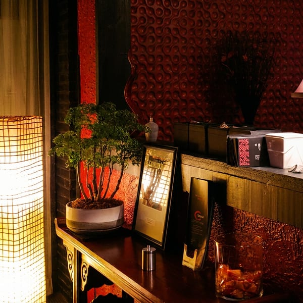 Warmly lit restaurant interior with glowing shoji screens, red textured walls, decorative lamps, framed menus, and a potted plant.